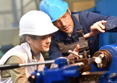 An Asian woman in a white hard hat receiving training on manufacturing equipment from a Caucasian male in a teal blue hard hat.
