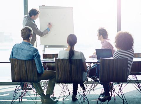 Group of business people sitting at a training room conference table looking at man at easel.